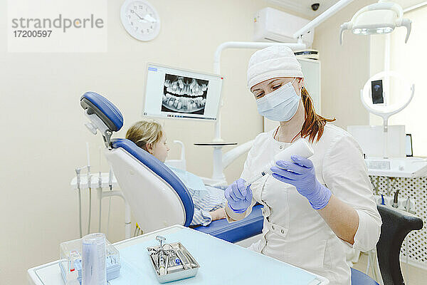 Zahnärztin mit Gesichtsschutzmaske bereitet Spritze für Injektion vor  im Hintergrund sitzt ein kleines Mädchen