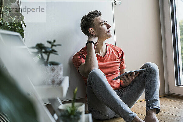 Nachdenklicher junger Mann mit digitalem Tablet  der wegschaut  während er im Wohnzimmer sitzt