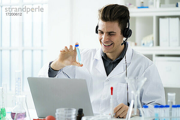 Lächelnde junge Wissenschaftlerin  die ein Reagenzglas während eines Videogesprächs auf einem Laptop im Labor zeigt