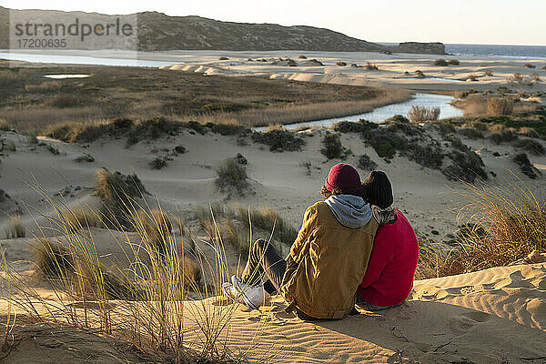 Paar mit Strickmütze sitzt bei Sonnenuntergang auf einer Sanddüne zusammen