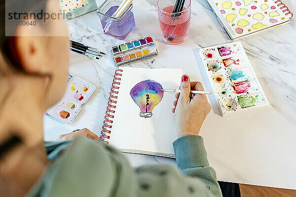 Frau malt Glühbirne mit Aquarellfarben auf Spiralnotizbuch