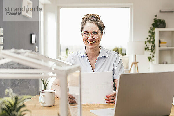 Fröhliche Frau mit Brille  die ein Dokument hält  während sie am Schreibtisch sitzt