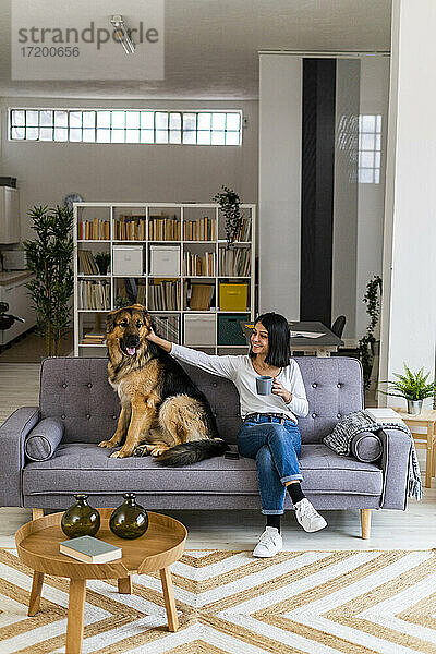 Junge Frau streichelt ihren Hund  während sie auf dem Sofa im Wohnzimmer sitzt