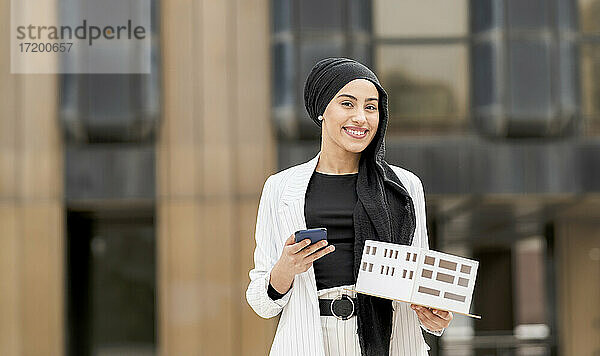 Lächelnde Architektin mit Smartphone hält Architekturmodell im Freien