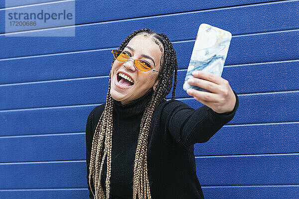 Glückliche Frau  die ein Selfie mit ihrem Mobiltelefon macht  während sie an einer blauen Wand steht