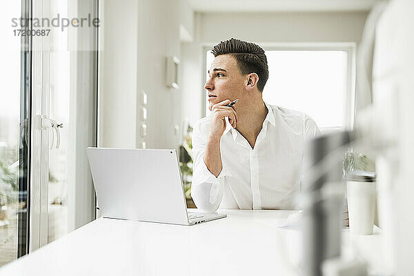 Junger männlicher Berufstätiger  der durch das Fenster schaut  während er in seinem Büro zu Hause sitzt