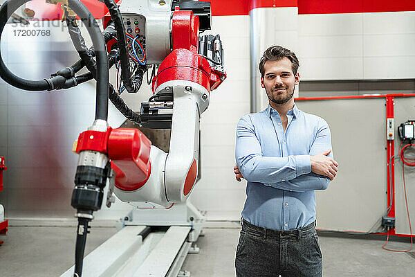 Männlicher Ingenieur mit verschränkten Armen an einem Roboter in einer Fabrik stehend