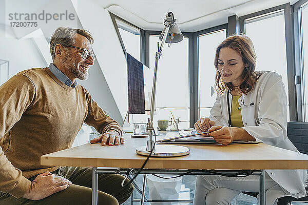 Lächelnder Arzt  der ein digitales Tablet benutzt  während er neben einem Patienten in der Klinik sitzt