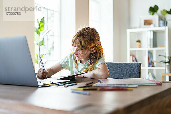 Konzentriertes rothaariges Mädchen macht Hausaufgaben vor dem Laptop zu Hause