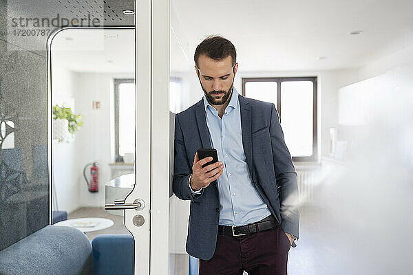 Junger Unternehmer  der ein Mobiltelefon benutzt  während er sich an eine Telefonzelle im Büro lehnt