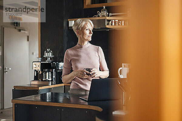 Lächelnde Geschäftsfrau  die wegschaut  während sie eine Kaffeetasse in der Küche ihres Heimbüros hält
