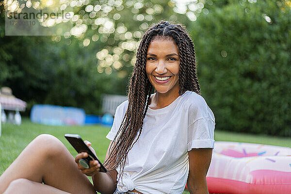 Lächelnde  geflochtene Frau mit Handy in der Hand  die im Garten sitzt