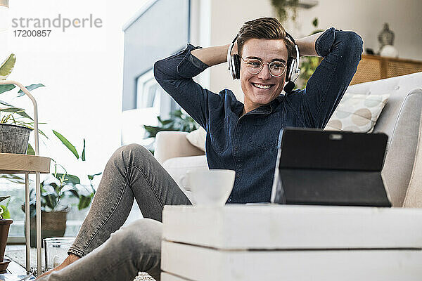 Glücklicher junger Mann mit drahtlosen Kopfhörern  der im Wohnzimmer ein Video über ein digitales Tablet ansieht