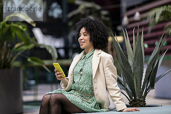 Nachdenkliche afroamerikanische Frau mit Smartphone sitzt an einer Topfpflanze