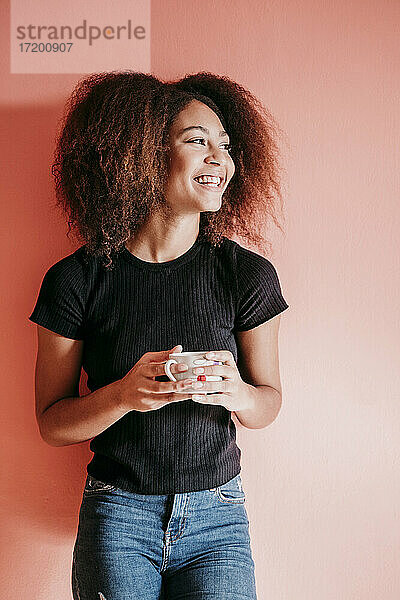 Glückliche Afro-Frau  die eine Kaffeetasse hält und vor einem pfirsichfarbenen Hintergrund wegschaut