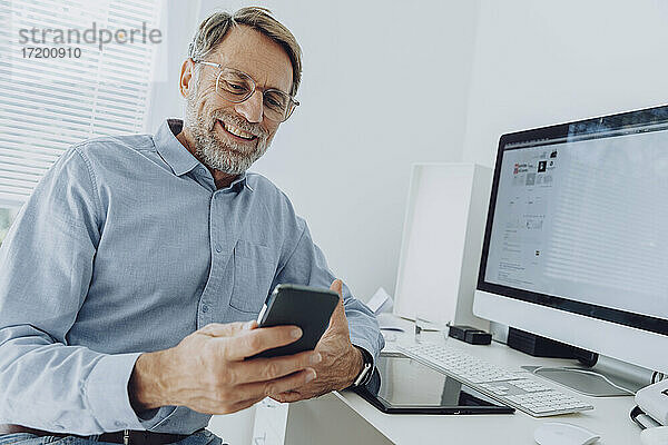 Lächelnder reifer Geschäftsmann mit Brille  der im Büro zu Hause ein Mobiltelefon benutzt