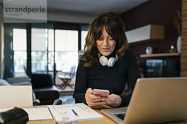 Frau mit Laptop  die lächelt  während sie ein Mobiltelefon zu Hause benutzt