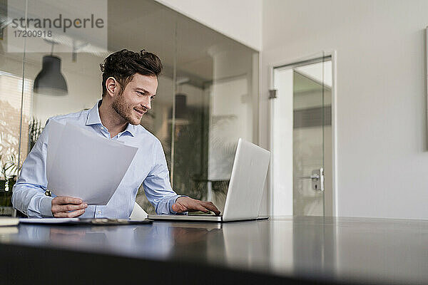 Lächelnder männlicher Unternehmer mit Dokument  der im Büro an einem Laptop arbeitet