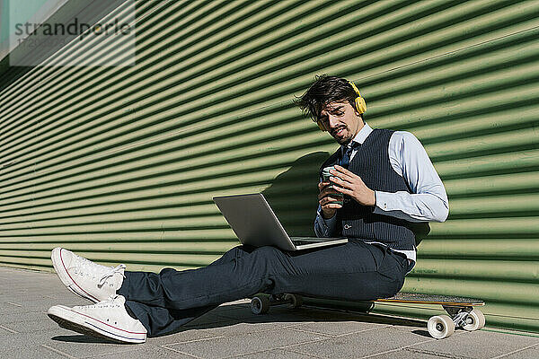 Geschäftsmann mit Laptop  der eine Kaffeetasse hält und auf einem Longboard vor einer grünen Wand sitzt