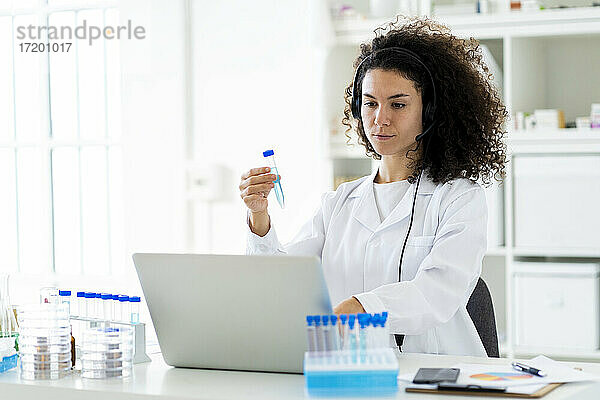 Wissenschaftlerin mit Reagenzglas bei der Arbeit am Laptop in einer Klinik