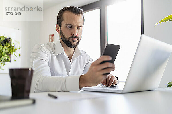 Junger Unternehmer mit Laptop  der ein Mobiltelefon benutzt  während er im Büro sitzt