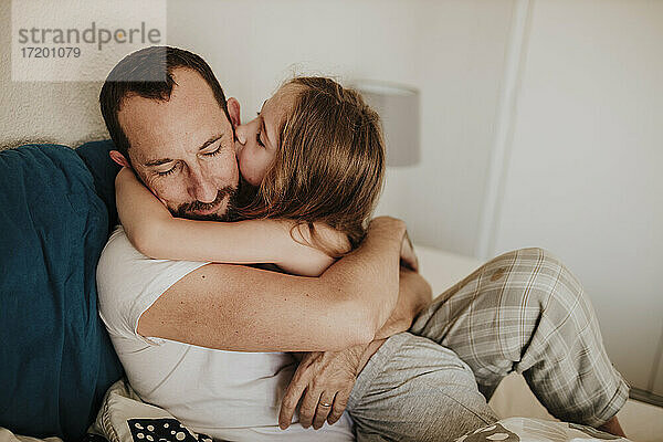 Tochter küsst Vater auf die Wange beim Umarmen auf dem Bett zu Hause