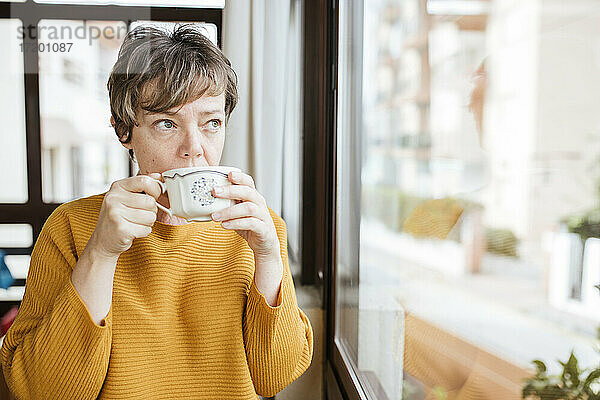 Reife Frau trinkt Kaffee und schaut durch das Fenster zu Hause