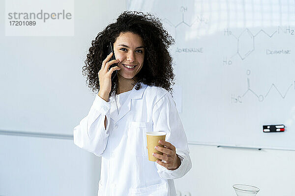 Lächelnde Ärztin mit Kaffeetasse  die in einer Klinik mit einem Mobiltelefon spricht