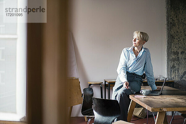 Lächelnde Geschäftsfrau mit Laptop auf dem Schreibtisch sitzend  während sie im Büro zu Hause wegschaut