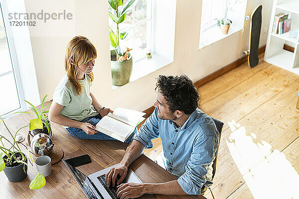 Rothaariges Mädchen mit Buch schaut auf Vater  der von zu Hause aus am Laptop arbeitet