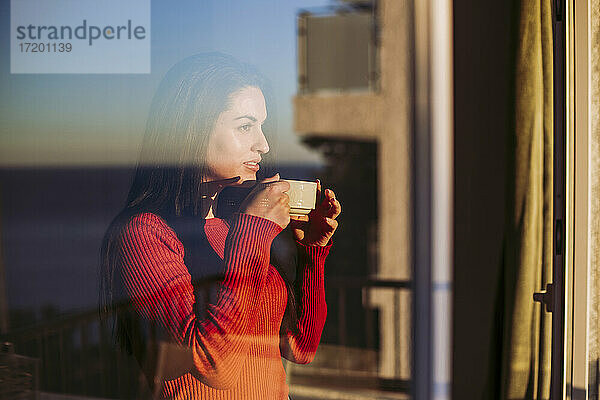 Nachdenkliche Frau  die einen Kaffee trinkt  während sie durch ein Fenster zu Hause schaut