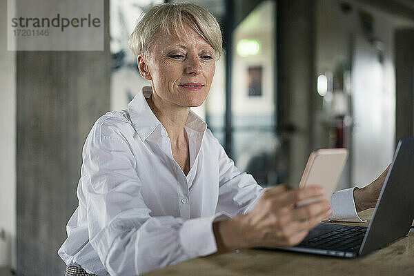 Blonde Geschäftsfrau mit Laptop und Mobiltelefon am Schreibtisch im Heimbüro
