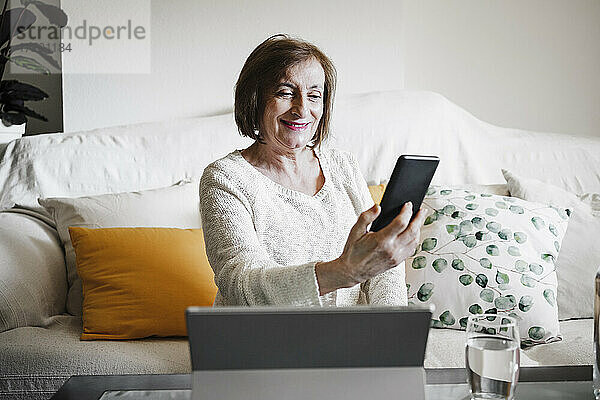 Lächelnde Frau  die ein Mobiltelefon benutzt  während sie zu Hause auf dem Sofa sitzt