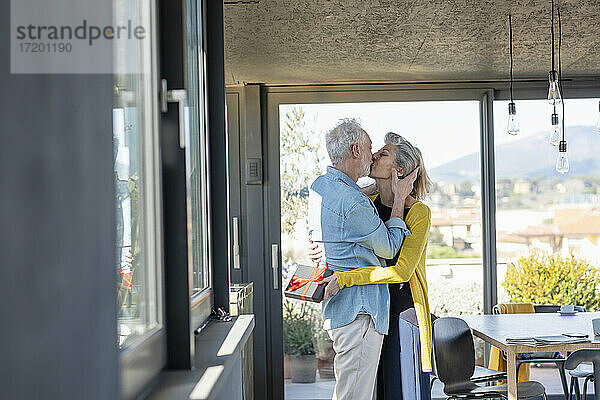 Ältere Frau mit Geschenkbox  die einen Mann küsst  während sie zu Hause steht