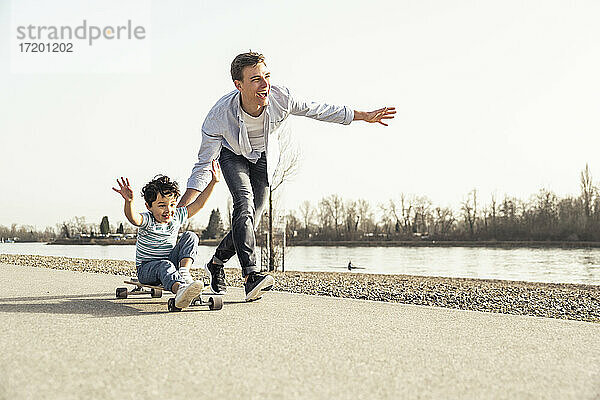 Vater läuft und schiebt seinen Sohn  der auf einem Skateboard sitzt  an einem sonnigen Tag