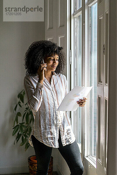 Weibliche Afro-Unternehmerin mit Strategie im Gespräch auf dem Smartphone  während sie am Fenster im Büro steht