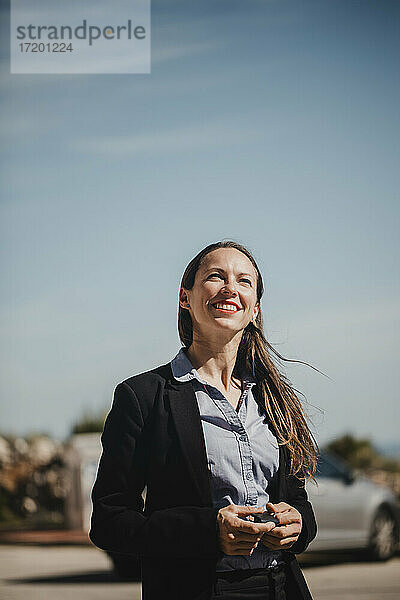 Lächelnde Geschäftsfrau mit Blick nach oben  während sie an einem sonnigen Tag gegen den Himmel steht