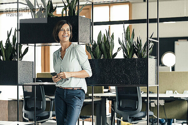 Lächelnde Geschäftsfrau mit Smartphone  die wegschaut  während sie im Büro steht