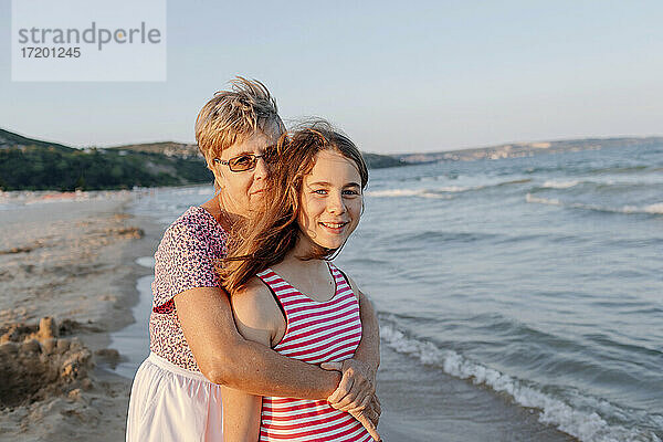 Ältere Frau umarmt ihre Enkelin  während sie am Strand steht