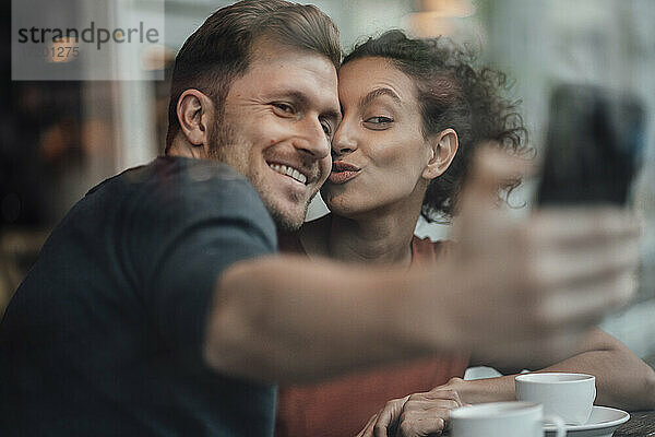 Lächelnder Freund nimmt Selfie mit Freundin durch Handy beim Sitzen im Café