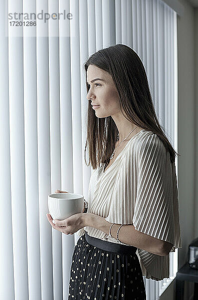 Junge Frau mit Kaffeetasse  die wegschaut  während sie zu Hause am Fenster steht