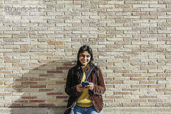 Lächelnde Frau mit Umhängetasche  die ihr Mobiltelefon gegen eine Backsteinmauer hält