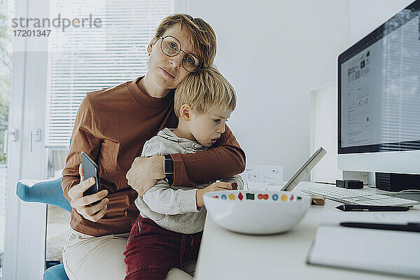 Liebevolle Mutter  die ihren Sohn umarmt  während sie im Büro zu Hause auf ein digitales Tablet schaut