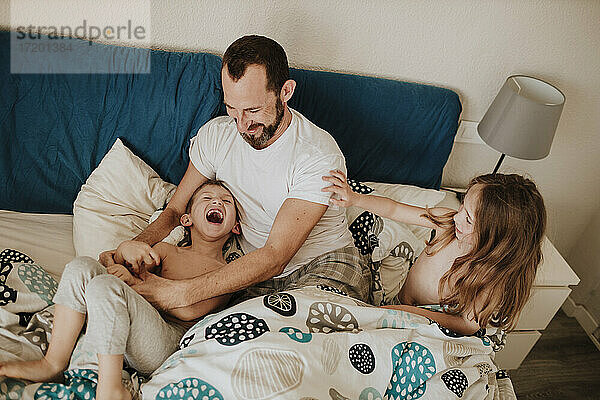 Vater spielt mit fröhlichem Sohn  während Tochter zu Hause auf dem Bett sitzt