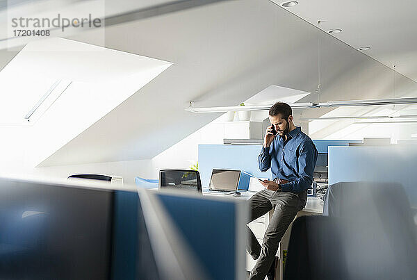 Junger Geschäftsmann  der ein digitales Tablet benutzt  während er im Büro mit einem Mobiltelefon spricht