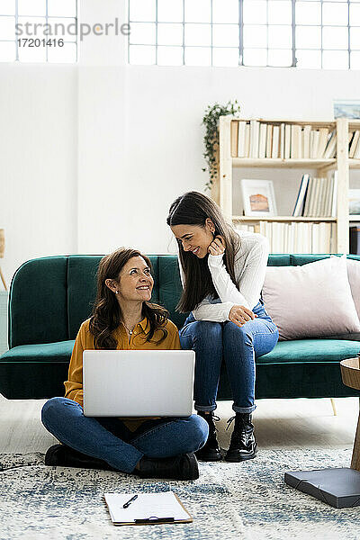 Mutter mit Laptop lächelt Tochter an  während sie auf dem Sofa im Wohnzimmer sitzt