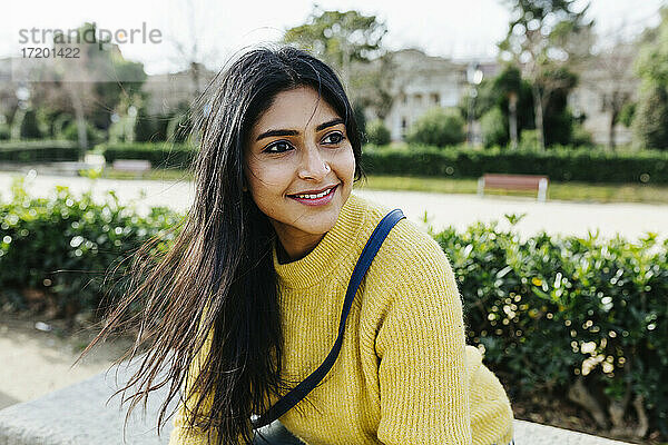 Lächelnde Frau im Pullover  die wegschaut  während sie im Park sitzt