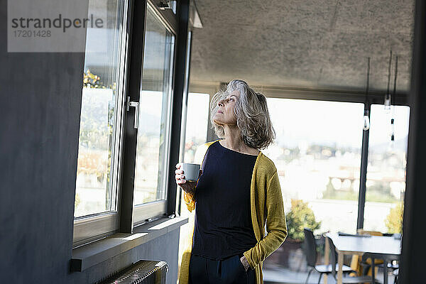 Nachdenkliche Frau mit Kaffeetasse  die zu Hause am Fenster steht