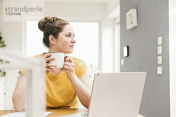 Unternehmerin hält Kaffeetasse  während sie am Schreibtisch sitzt und wegschaut