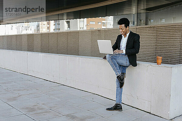 Männlicher Unternehmer  der einen Laptop benutzt  während er auf einem Bein gegen eine Stützmauer steht